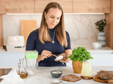 Mujer untando queso de kéfir en una rebanada de pan en una cocina, con albahaca y otros ingredientes frescos.