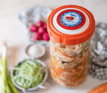 Un tarro de vidrio Kefirko con tapa naranja y vegetales en escabeche reposa junto a productos frescos en la cocina.