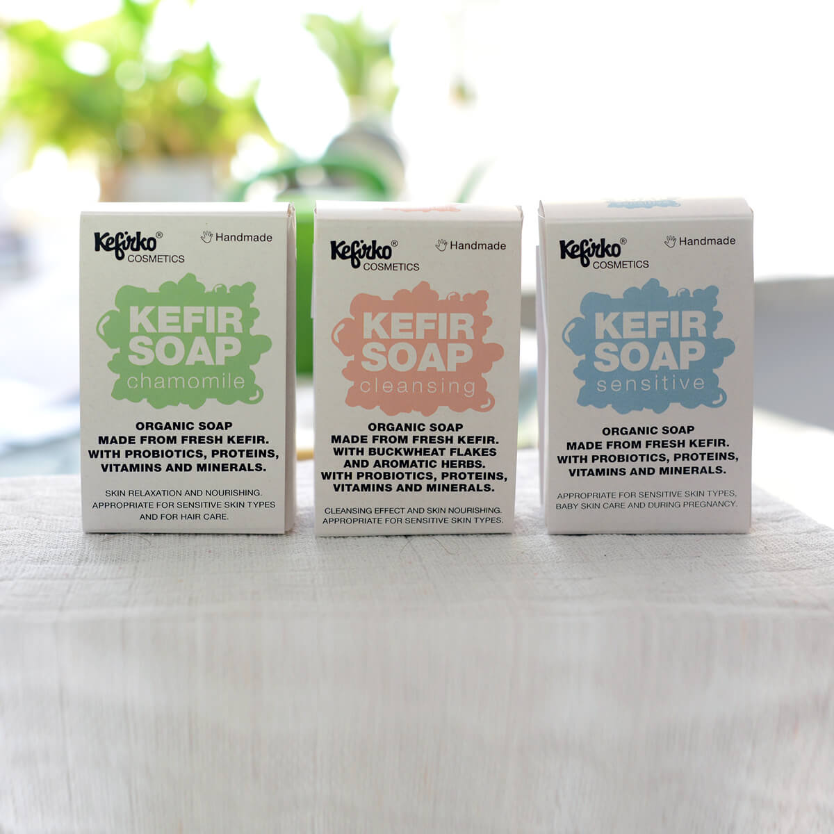 Tres cajas de jabones probióticos Kefirko (manzanilla, limpiador, sensible) se colocan sobre una superficie de madera con vegetación borrosa de fondo.