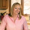 Donna Schwenk con una camisa rosa sonriendo mientras está de pie en una cocina con gabinetes de madera y una ventana al fondo, sosteniendo un recipiente de vidrio lleno de kéfir.