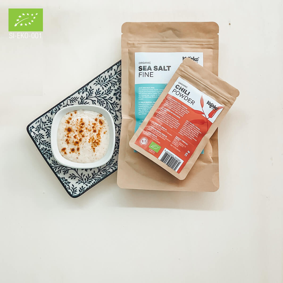 Un recipiente que contiene un plato cremoso se encuentra junto a paquetes etiquetados como ingredientes esenciales, que incluyen sal marina y chile en polvo, en un plato decorativo.