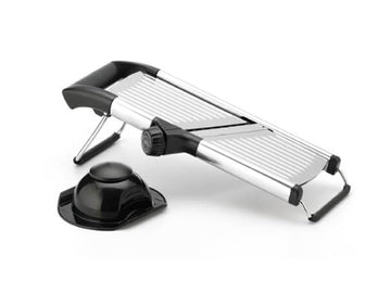 Una mandolina cortadora de acero inoxidable con una cuchilla ajustable y un soporte de seguridad negro a su lado, junto a un recipiente de vidrio lleno de kéfir.