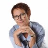 Suzana Kranjec con el pelo corto y gafas, vestida con una camisa de rayas azules, se inclina hacia delante con las manos entrelazadas bajo la barbilla y sonríe. Frente a ella hay un recipiente de cristal que contiene kéfir.