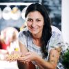 Urska fartelj está parada en la cocina, con su cabello largo cayendo en cascada mientras sonríe alegremente, sosteniendo un puñado de pasta en su mano. Junto a ella está el producto Urska de Kefirko.