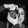 Verdana Orlovic sonríe y sostiene una cámara frente a su rostro, en blanco y negro. La lente de la cámara está centrada en el ojo y hay un recipiente de vidrio con kéfir sobre la mesa frente a ella.