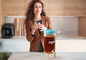 Una mujer sonriente con americana marrón sostiene un vaso de kombucha junto a un gran fermentador Kefirko en una cocina moderna.