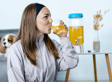 Mujer bebiendo de un vaso. Al fondo, un dispensador con una bebida probiótica y un perro en un sofá.