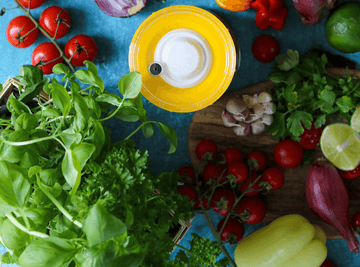 Vista cenital de un tarro de fermentación rodeado de verduras de primavera frescas como tomates, hierbas y cebollas.