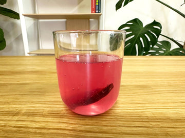 Vaso de vidrio con un kéfir tónico rosado burbujeante y un trozo de remolacha en su interior, sobre una mesa de madera.