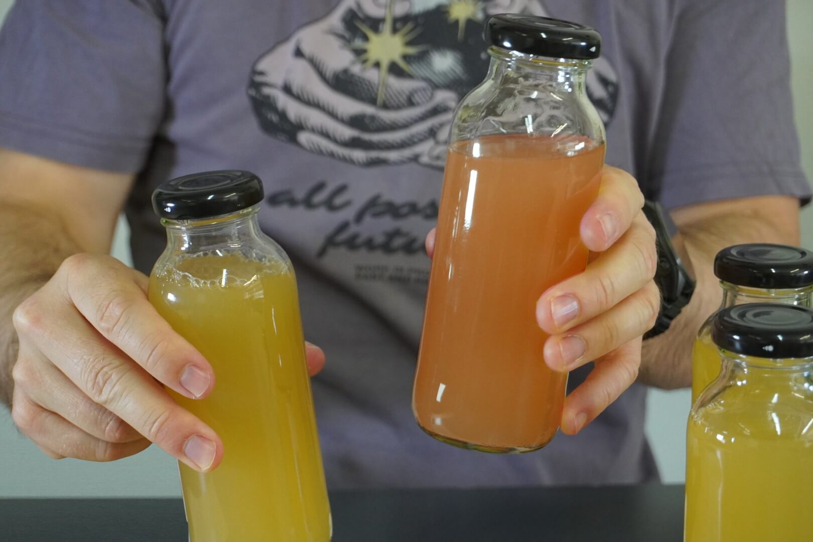Una persona sostiene dos botellas de vidrio Kefirko con bebidas amarillas y rosas, con una camiseta morada con un diseño gráfico.