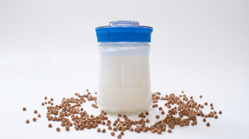 Tarro de vidrio Kefirko con kéfir de leche, rodeado de granos de trigo sarraceno sobre una superficie blanca.