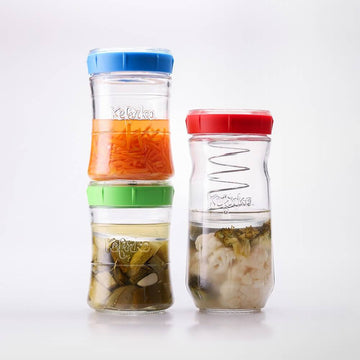 Tres tarros de vidrio Kefirko con tapas de colores llenos de diferentes alimentos fermentados como zanahorias y pepinillos.