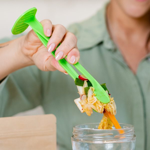 Mano usando unas pinzas verdes para sacar una mezcla de verduras de un tarro de vidrio, preparación de fermentación casera.