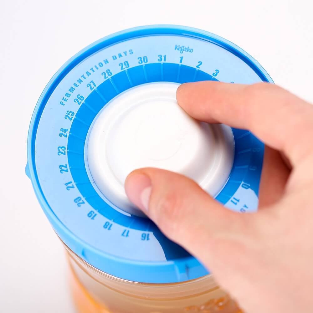 Mano ajustando el dial contador de días en la tapa azul del tarro de fermentación Kefirko.
