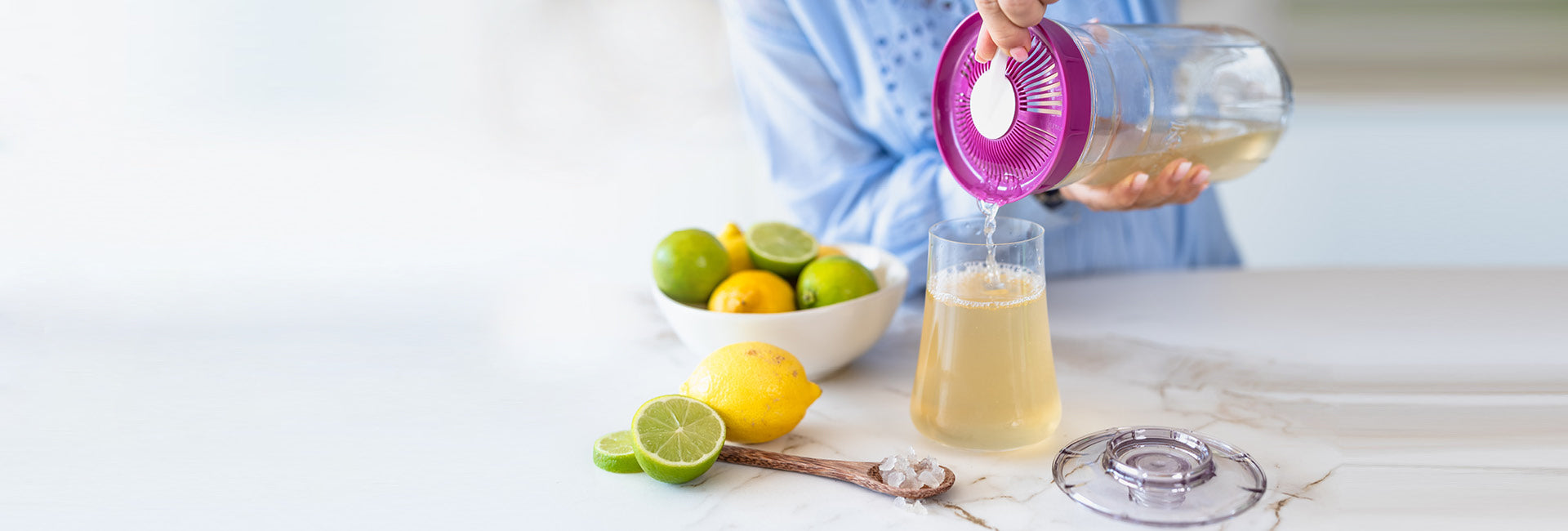 Una persona vierte limonada refrescante de una jarra en un vaso sobre una mesa al lado de un recipiente con limones y limas picantes, todo ello colocado al lado del fermentador de kéfir Kefirko que contiene un líquido blanco, lo que indica que podría ser kéfir de leche.slider_item_4KBqeF
