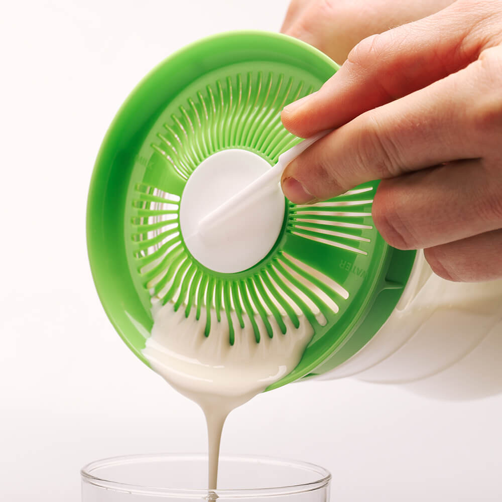 Una mano vierte kefir de leche espeso a través de la tapa colador del fermentador Kefirko en un vaso sobre un fondo blanco.