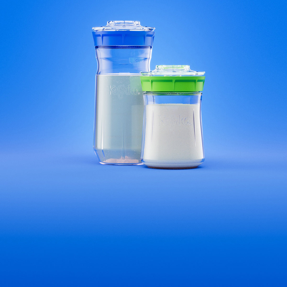 Dos fermentadores Kefirko con cuerpos transparentes y tapas verdes y azules sobre un fondo azul; el fermentador más grande contiene un líquido blanco, mientras que el más pequeño contiene un polvo blanco.
