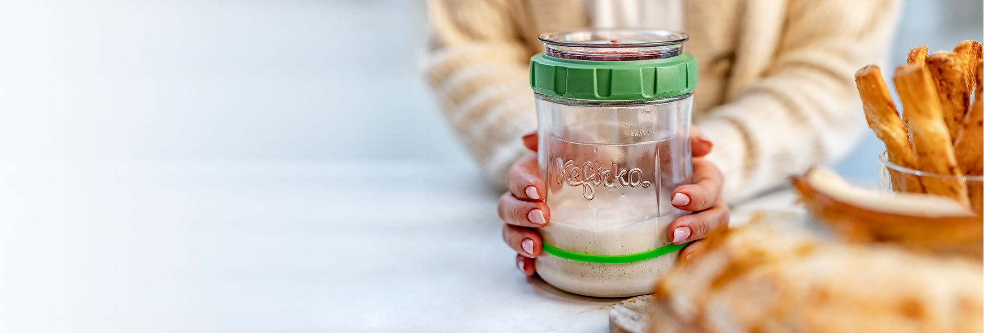 Manos sosteniendo un frasco de vidrio con una tapa verde etiquetada como "kefirko", diseñado como un fermentador de masa madre, adecuado para elaborar iniciadores de masa madre y otros productos fermentados.slider_item_keXmdg