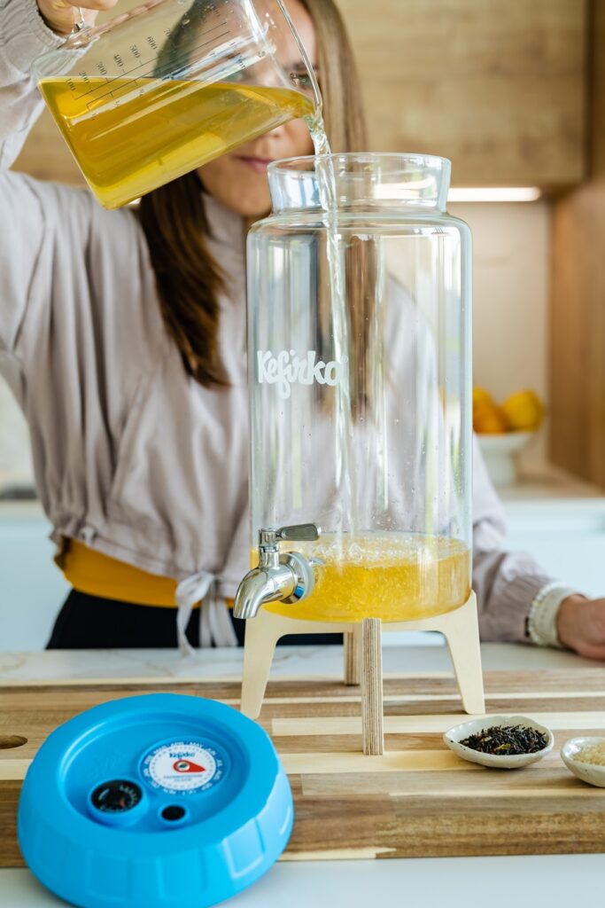 Persona vertiendo líquido de una jarra medidora en el tarro de vidrio Kefirko para hacer kombucha en una cocina.