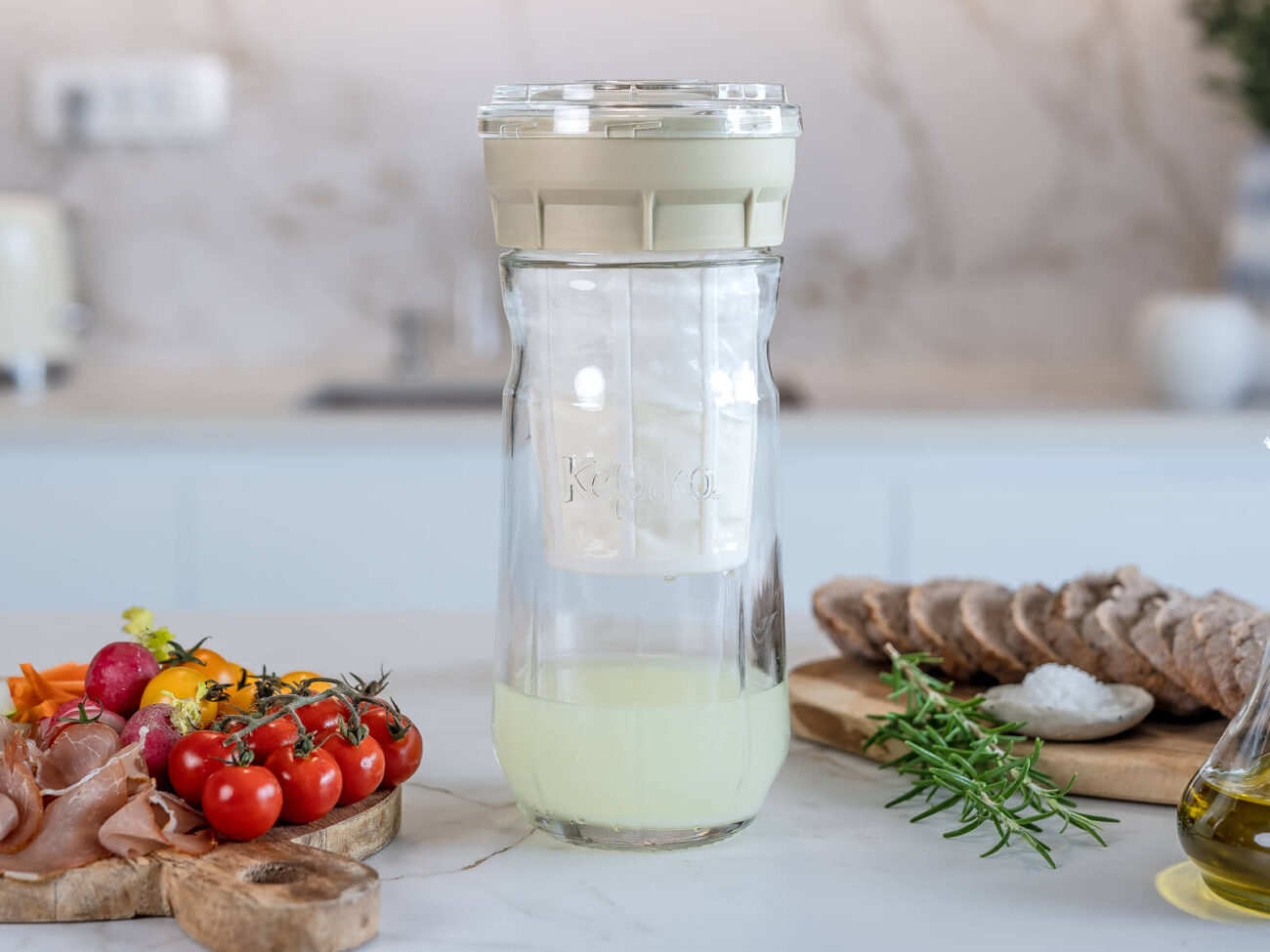 Bodegón mediterráneo con el fermentador de queso de Kéfir Kefirko como pieza central, junto a pan, tomates y aceite de oliva.