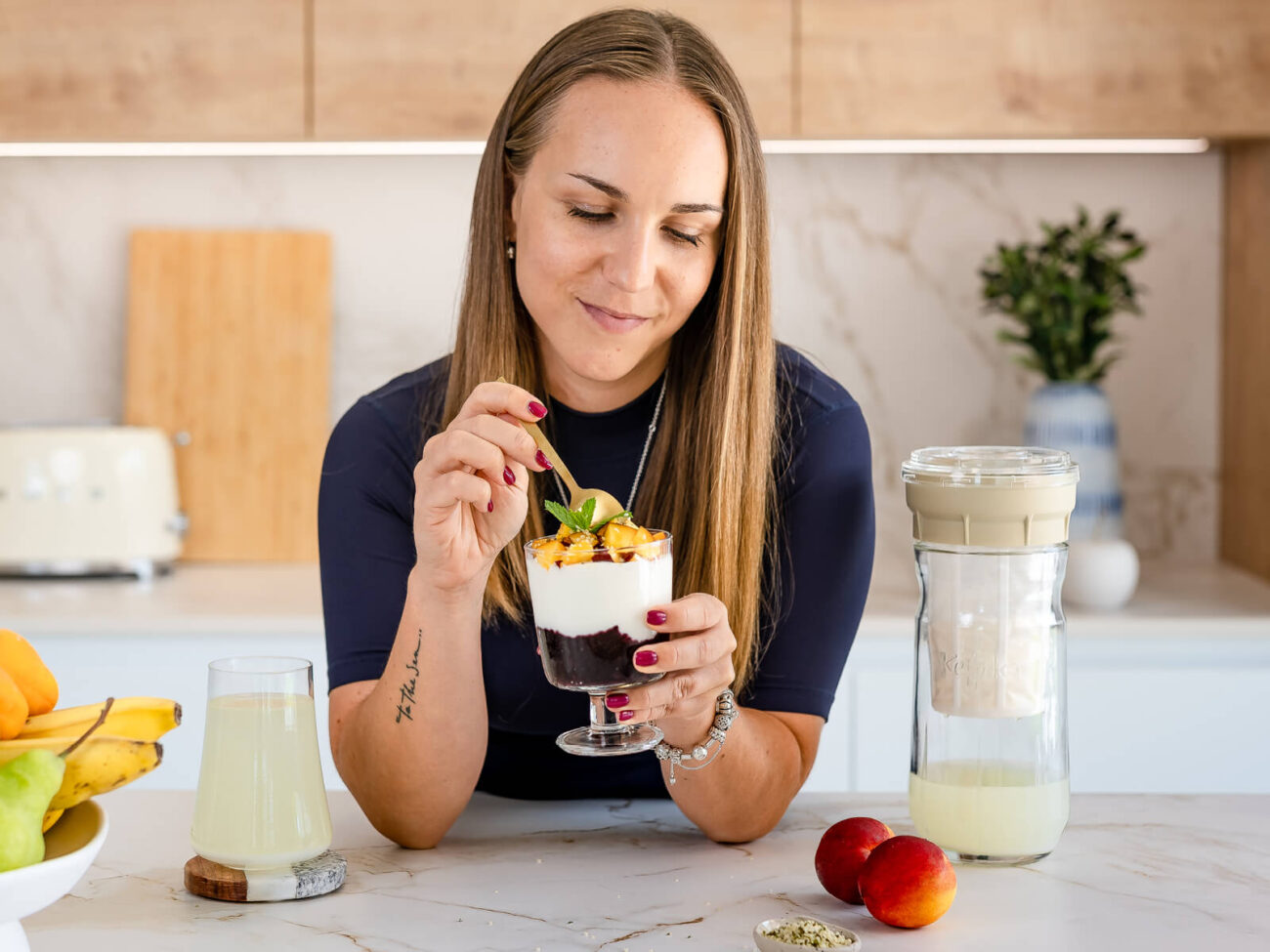 Escena de desayuno veraniego con una mujer disfrutando de un parfait y, al lado, el fermentador de queso de Kéfir Kefirko.