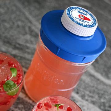 Botella para fermentación con una bebida rosada, junto a dos vasos servidos con kéfir y granada.